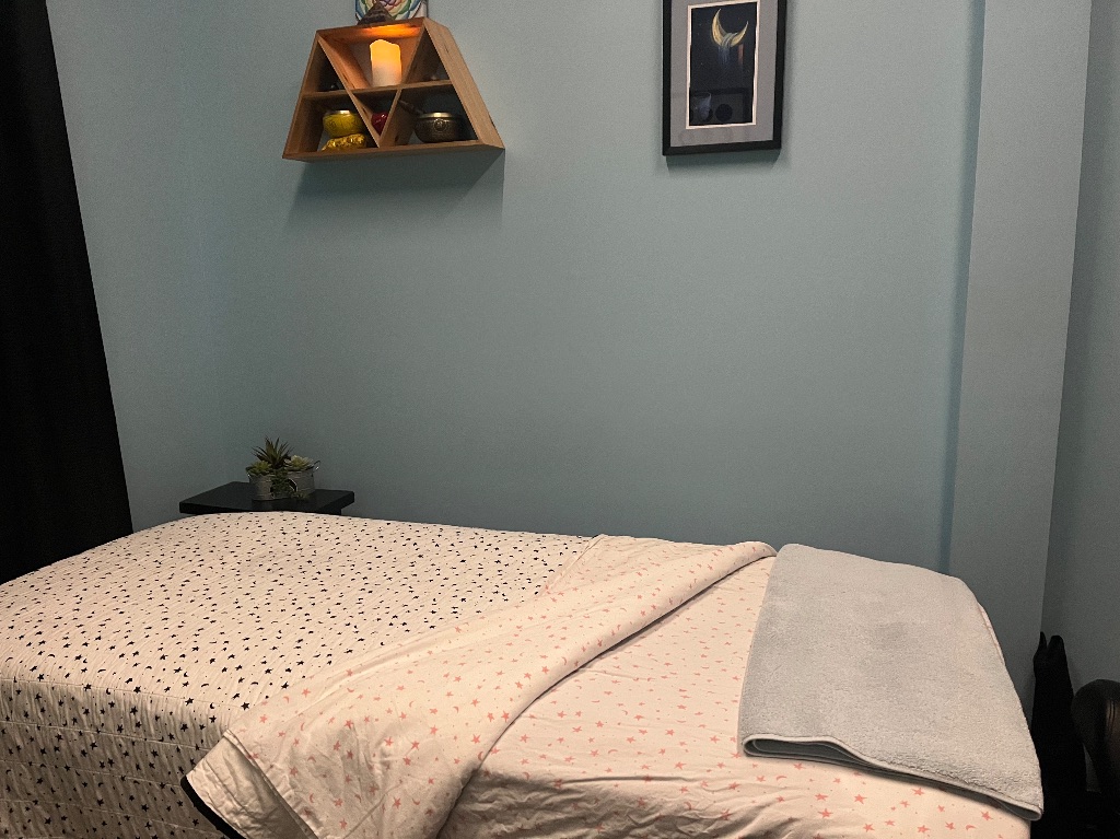 A massage table with moon sheets and towel sit in a small room with light blue walls there is a framed moon picture over it and a shelf with a candle on the wall. 