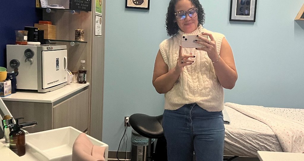 A brown woman with dark curly hair, a cream sleeveless sweater, and jeans stands in front of the mirror taking a picture with her phone. She is standing in front of a massage table against a light blue wall.