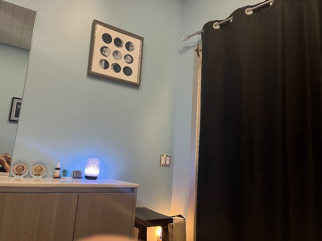 A small room with a black curtain on the right side. On the light blue wall is a framed picture of the moon phases and in front of a mirror is a dresser with a blue rock light on top. 