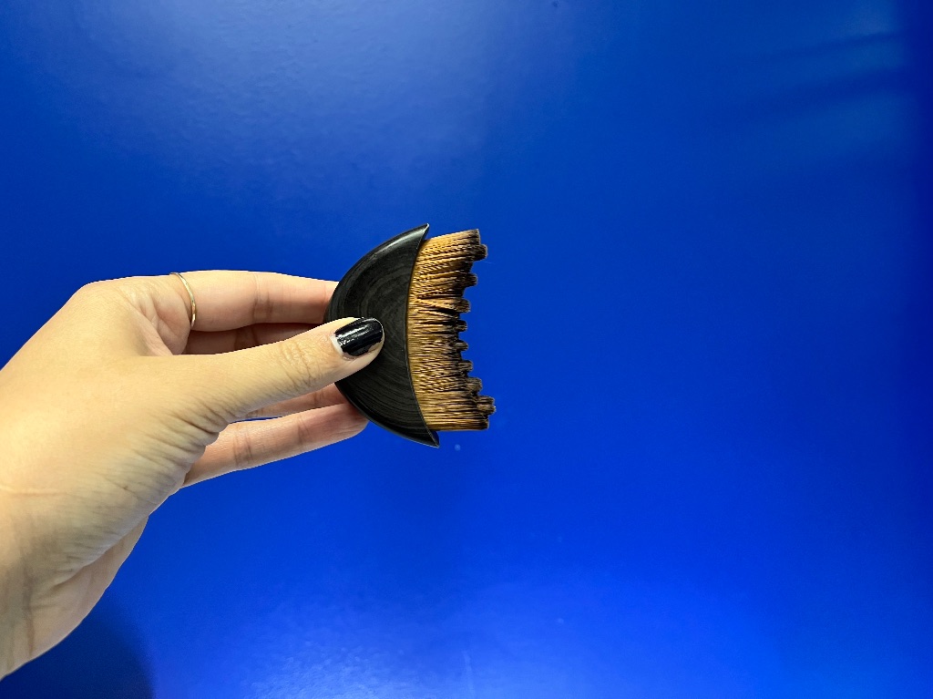A hand with dark nail polish holds a small half moon shaped brush with brown bristles, against a dark blue wall. 