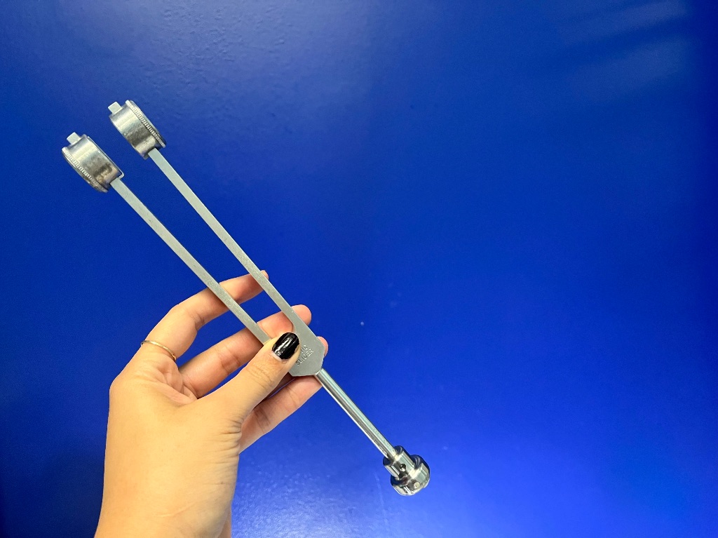 A hand with dark nail polish holds a metal tuning fork against a dark blue wall.