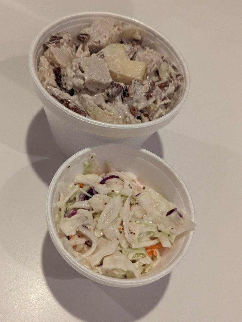 Two styrofoam containers sit on a table. The one in the back is bigger and filled with a turkey and apple pieces salad. The front container is full of creamy cole slaw