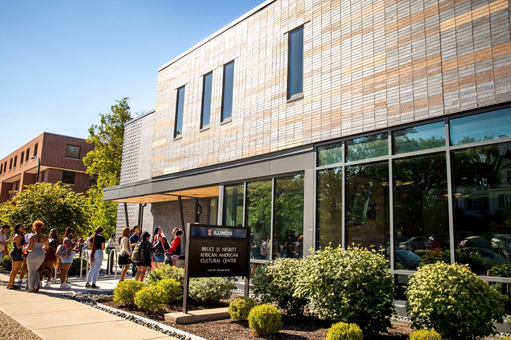The outside of the Bruce D. Nesbitt African American Cultural Center a stunning stone and glass two story building. There are small shrubs planted in front and a line of people waiting to enter the building on the left side of the image.