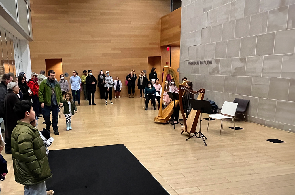 A crowd gathered in the lobby of a library watches a harpist perform.