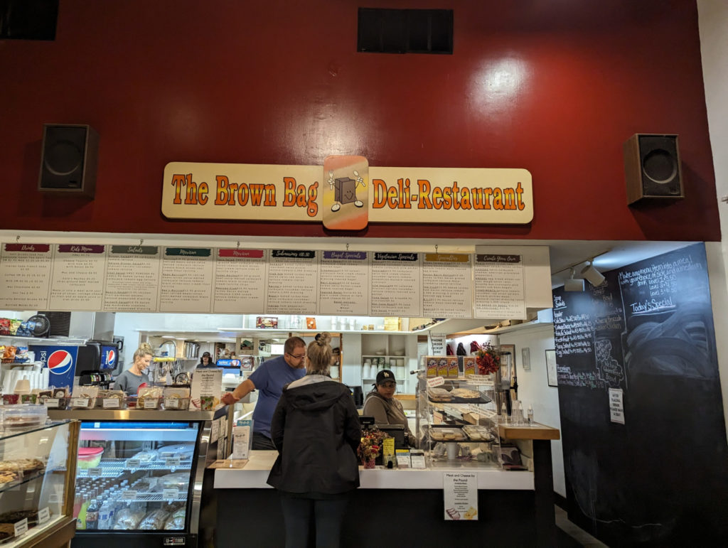 A large order counter with a menu board above. There is a cold deli case next to the counter and another pastry case on the left. The right wall is a black chalkboard and contains side options.