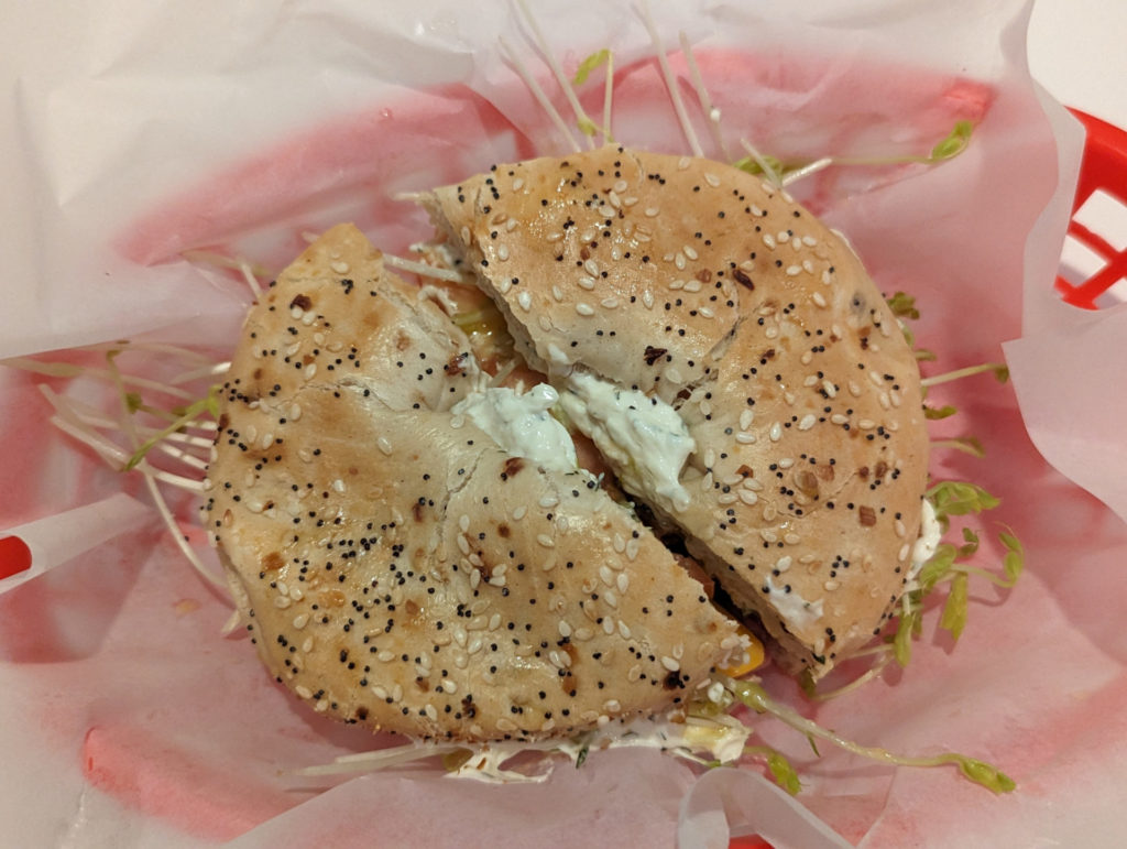 An everything bagel in a plastic tray with deli paper. Sprouts and cream cheese stick out from between the sandwich