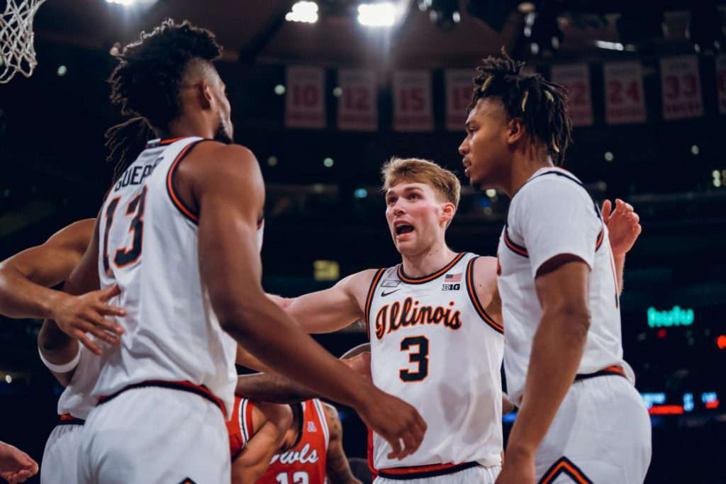 Four Illini men's basketball players huddle during a game. They are wearing white uniforms. Players are Marcus Domask (no. 3); Terrance Shannon, Jr. (pictured in profile), and Qunicy Guerrier (no. 13, pictured from behind). Another player is out of view from the camera, but an arm is around Guerrier.