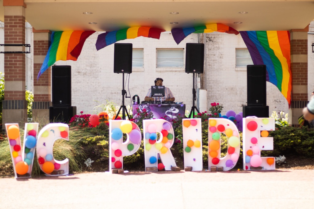 A stage with a DJ standing at a turntable between two large speakers. There are rainbow flags overhead and large balloon filled letters that say "VC PRIDE"