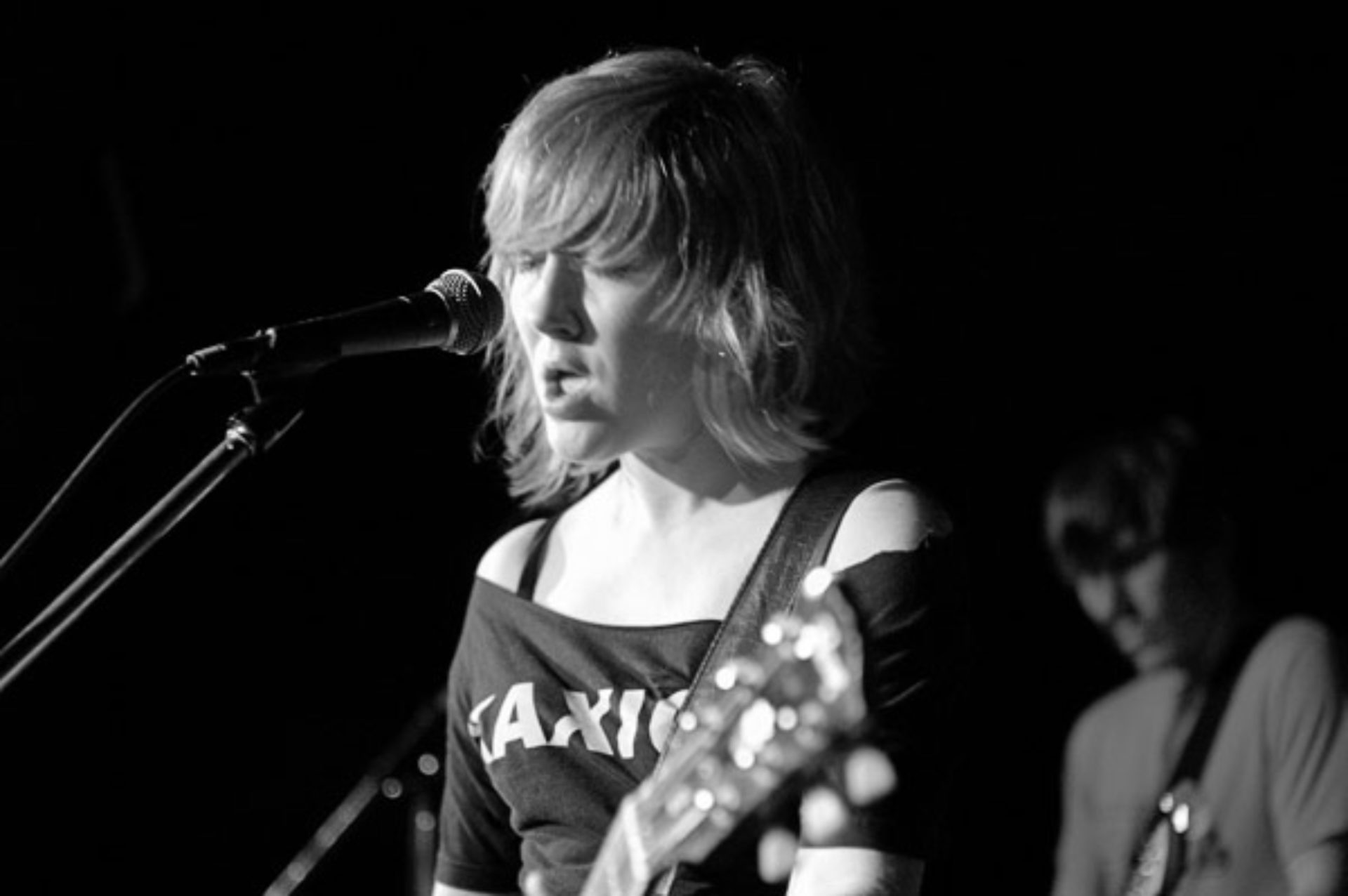 Sarge singer Elizabeth Elmore, a white woman, playing guitar and singing at a microphone. She has a bob with bangs, and is wearing what appears to be a cut up, collarless black t-shirt that lands below her shoulders.