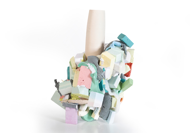 A ceramic sculpture with multiple light and pastel colored blocks of ceramic grouped and stuck to an ivory colored cylinder in the middle. The colorful pieces are adhered mostly to the bottom, with a slightly upward trajectory on the right side of the object, as pictured in this photo.