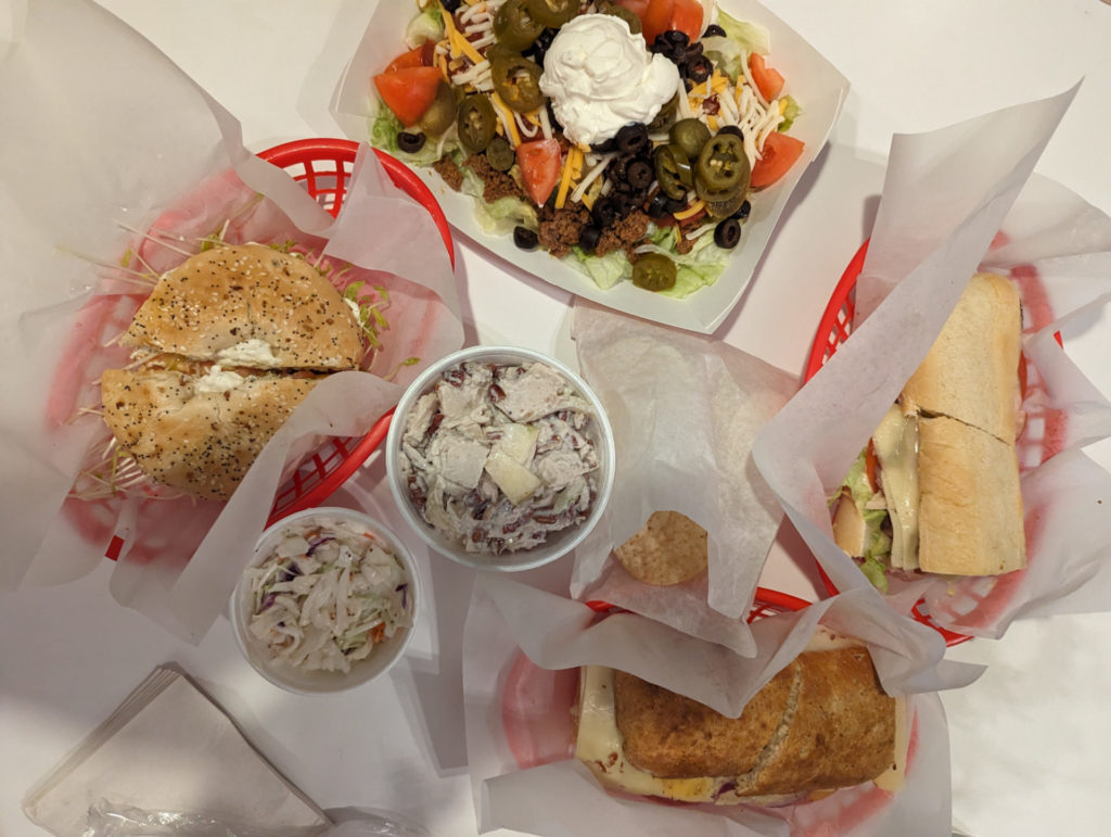 Taken from above, there are two sandwiches and a bagel plated on plastic trays with deli paper as well as a taco salad in a paper boat. In the middle are two sides; cole slaw and turkey salad. There is also a small bag of tortilla chips for the taco salad