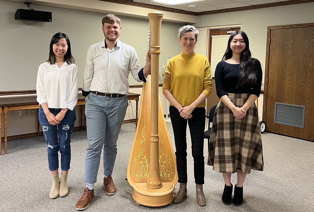 4 people stand next to a harp, 2 on each side. It appears to be in a room located in an educational institution.