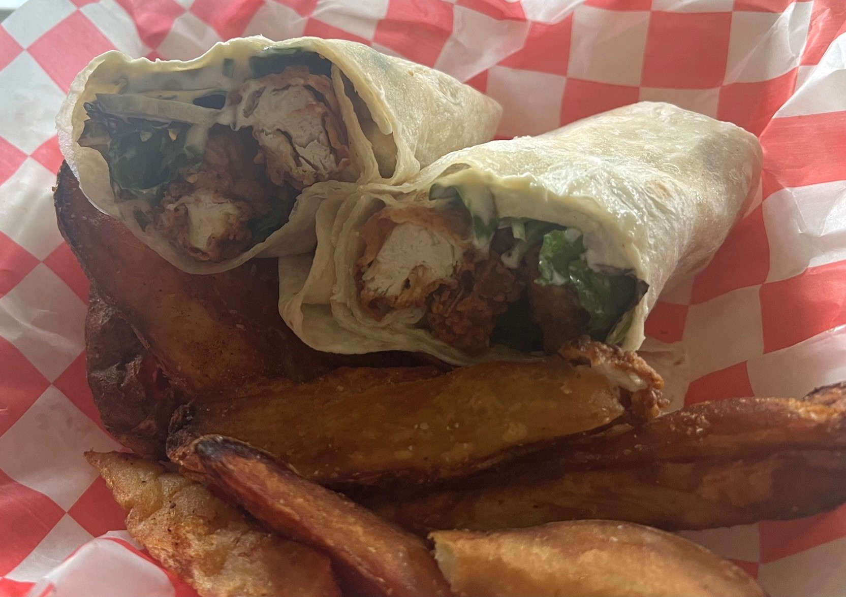 A fried chicken wrap, split in half, sitting in a container lined with red and white checked paper. There is a pile of potato wedges next to the wrap.