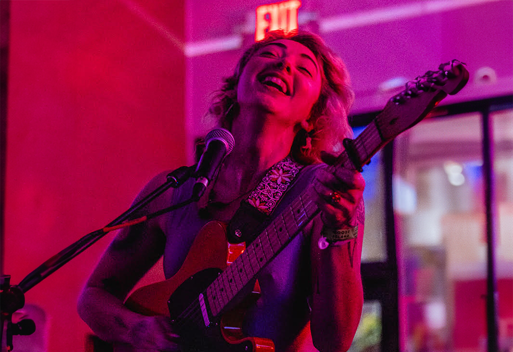 A performer onstage with red lighting playing a guitar and singing with their eyes closed