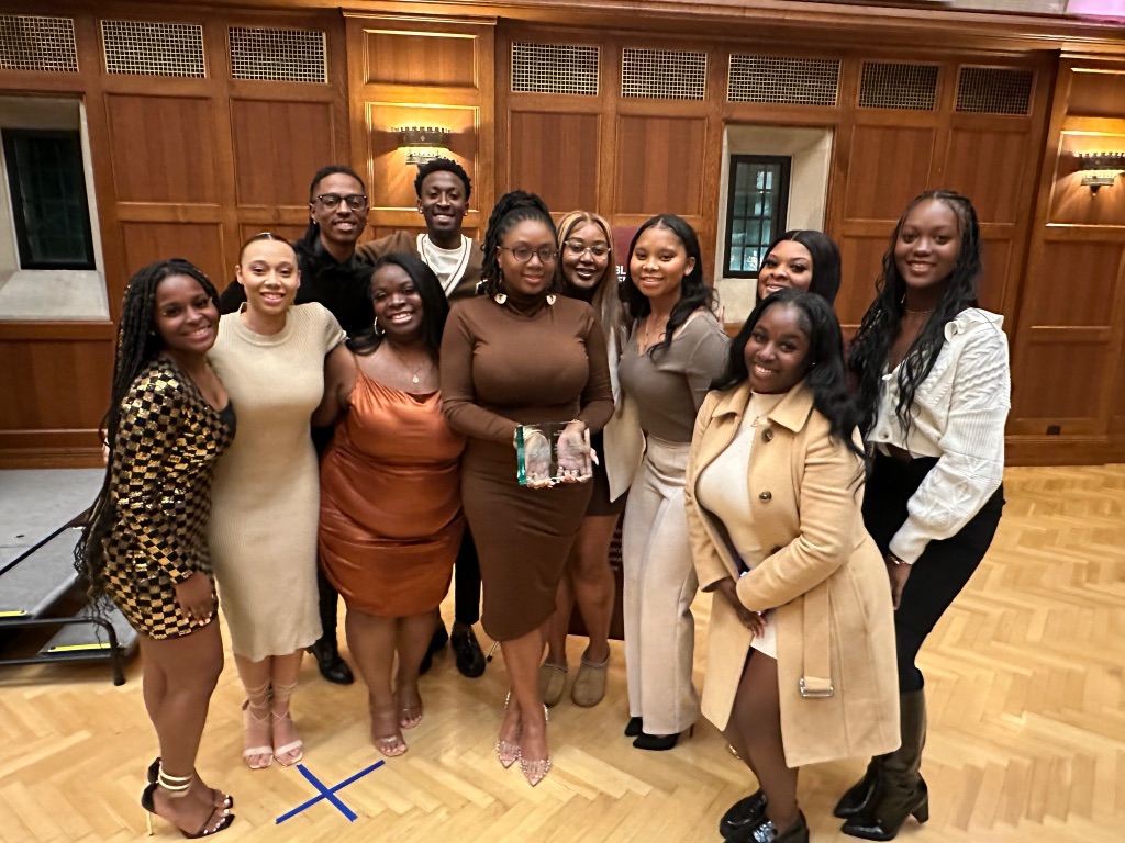 A group of black woman in brown, tan, and gold outfits surround a woman in a brown dress holding a clear award. They are standing in a wood paneled room 