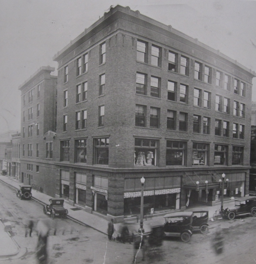 An old black and white photo of the Lincoln building from the early 1900's. The building is a 5 story brick building with big windows. There are old cars parked around it, and blurs of people walking by.