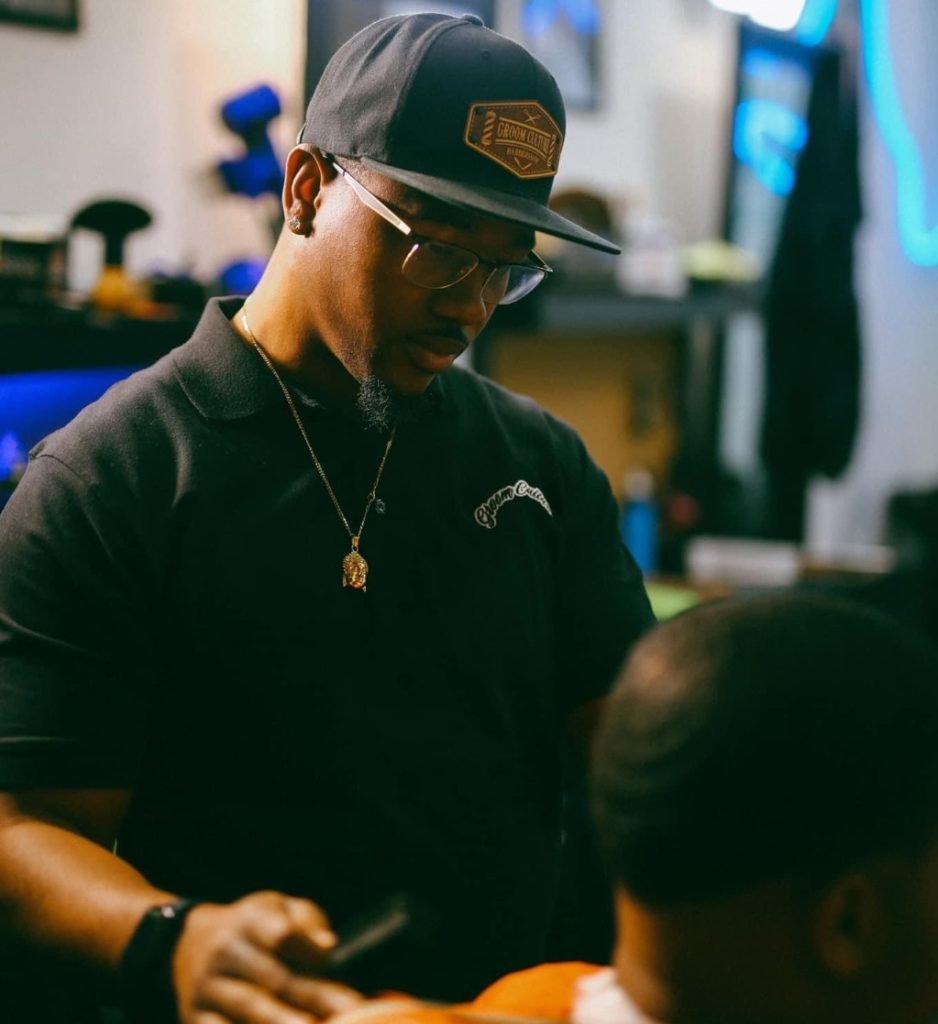 A black man in glasses, a black hat, t-shirt, and apron, standing next to another man about to cut his hair. 