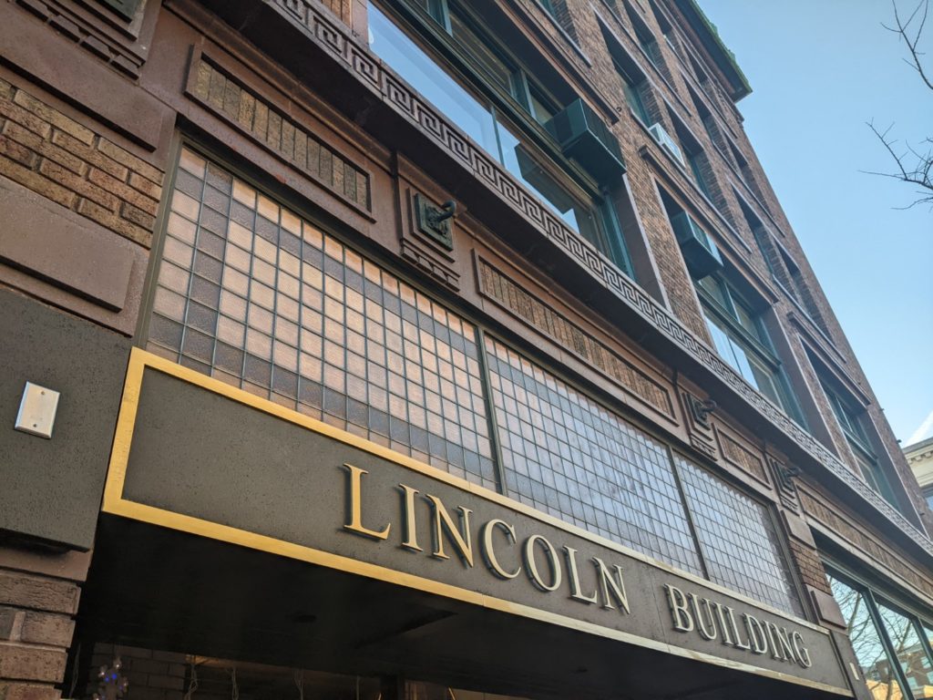 A close up of the Lincoln building. A big sign with the name is bronze colored letters and tiny panes of glass above and intricate metal work above.