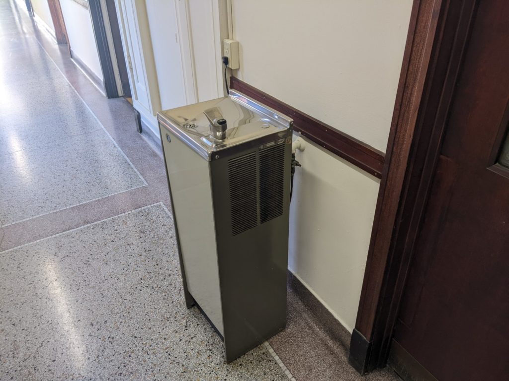 A gray water fountain in a hallway that plugs into the wall. 