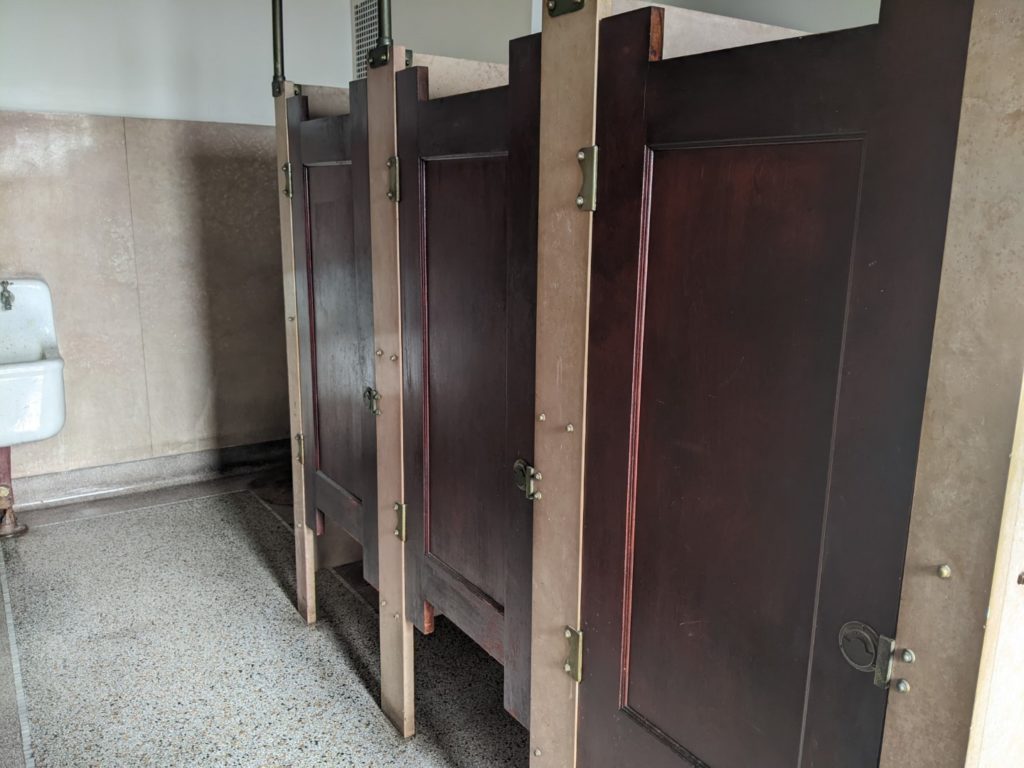 Bathroom marble stalls with wooden doors and brass finishes. There is a corner of a white sink visible on the left. 