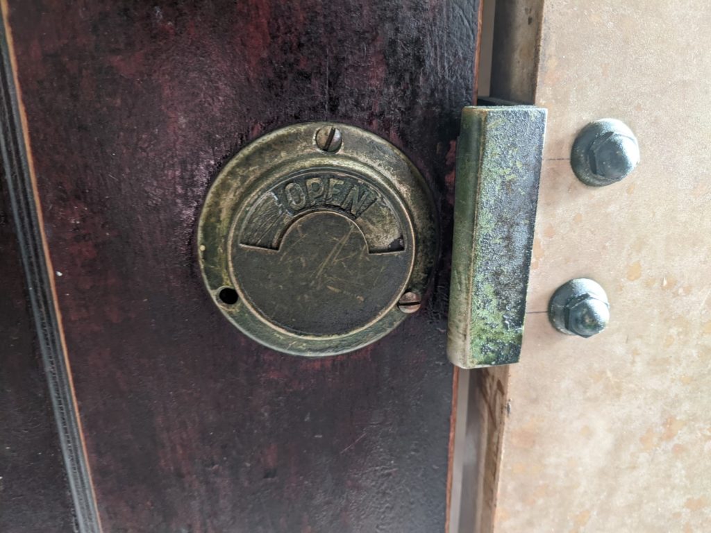 A close up shot of a green hinge and a brass bathroom stall lock. It is circular and has the words open visible