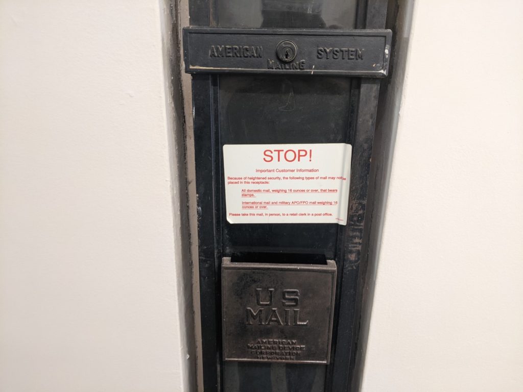 A black metal mail slot that has US MAIL stamped into the metal. There is a white sticker with red letters that say stop! 