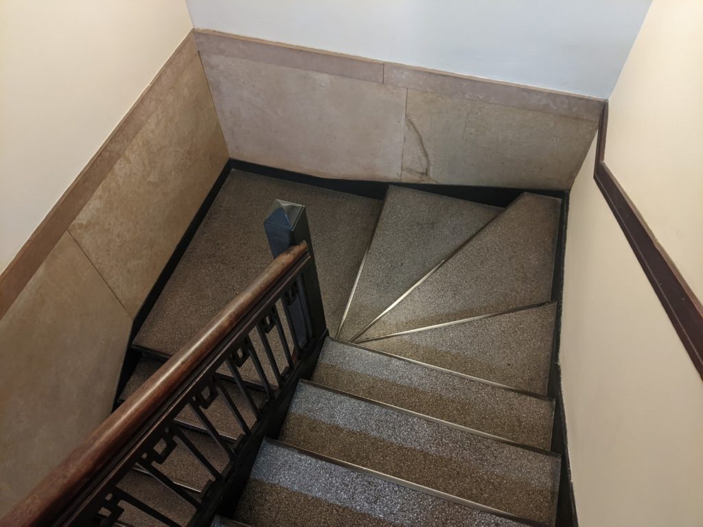 Looking down a steep staircase that curves around and goes down to the next floor. The steps are gray and the walls are white with a wooden bannister in the center. 