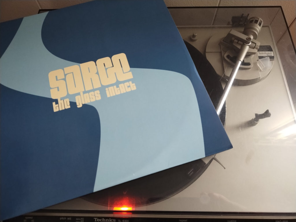 Vinyl album sleeve for Sarge's The Glass Intact. A dark blue background has a light blue river-like thick line vertically through the middle. Light yellow / cream colored block text reads "Sarge The Glass Intact." The album is resting on the lid of a turntable.