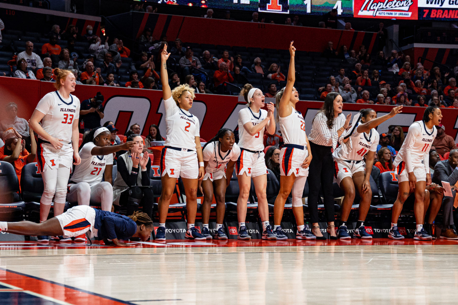 Illini women's basketball team sideline as viewed from the court. nine players are standing, squatting, or sitting and cheering on their teammakes. They are wearing white Illinois uniforms.