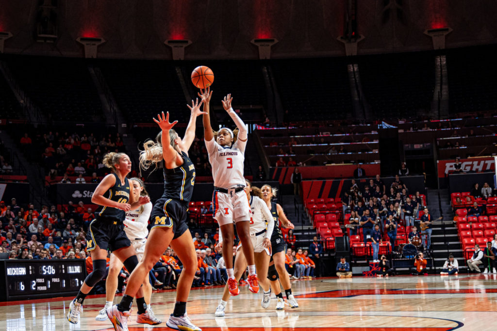 Illini women's basketball player Makira Cook, a Black woman wearing a white Illinois uniform, shoots a basketball during a game. She is being defended by a white woman in a Michigan uniform. The other players on the court are behind them.