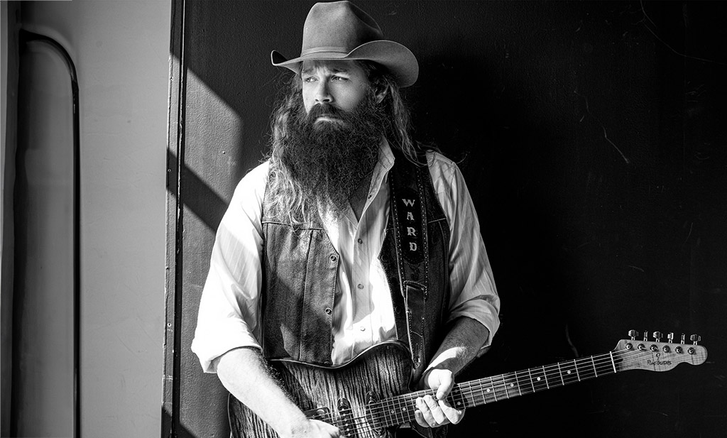 A black and white photo of a man in a cowboy hat holding a guitar looking out a window. His guitar strap says "Ward" on it.