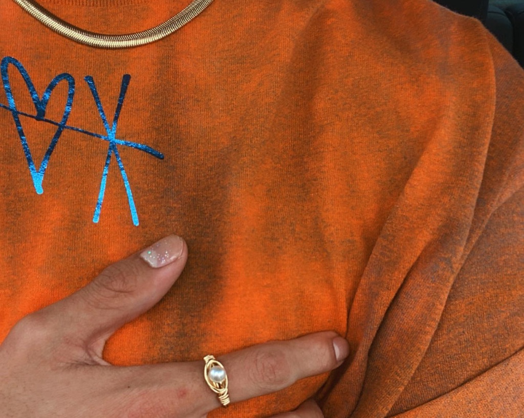 A close up of an orange t-shirt with a rainbow XO across the top center. There is a hand on the heart wearing a pear and gold ring. 