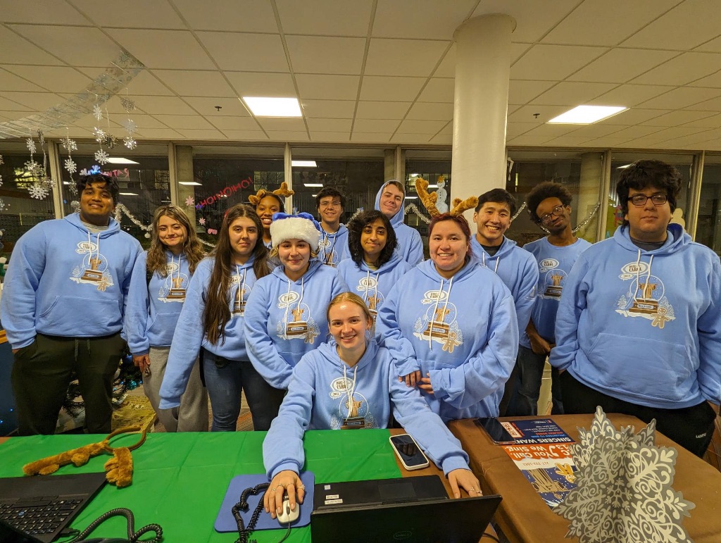 A group of students at a desk wear matching blue sweatshirts and sit in front of a desk with phones and computers.