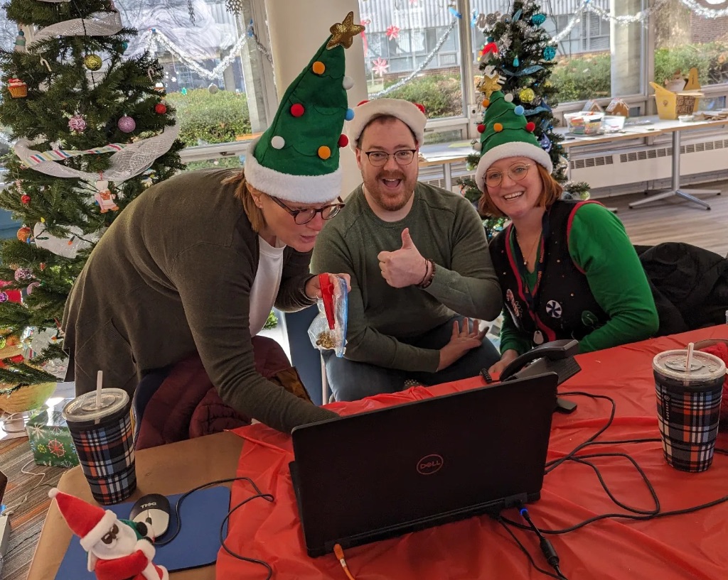 Three people sit in front of a computer screen wearing Santa hats and Christmas tree hats. 