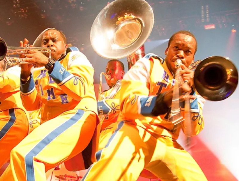 Two trumpet players and a tuba player in bright yellow marching band uniforms, playing their instruments in a crouched position.