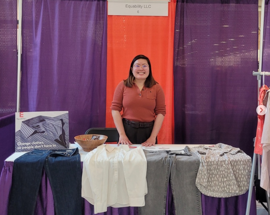 A woman with dark hair stands in front of a purple and orange curtain in front of a table with adaptive clothes.