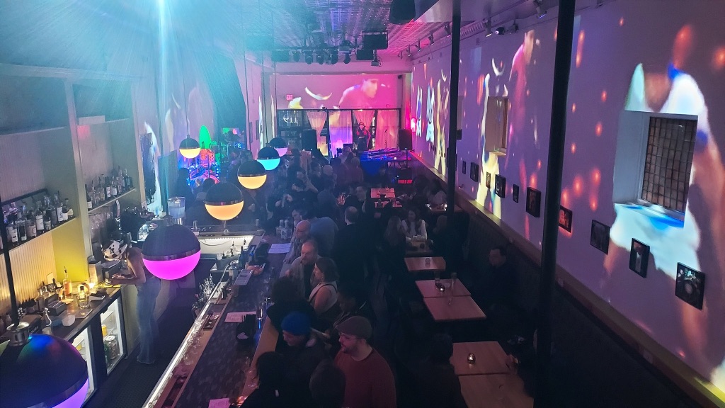 People crowd in the interior of Gallery Art Bar. has low tables on the right and bar seating on the left. Pendant lights are in shades of pink, purple, yellow, and green.