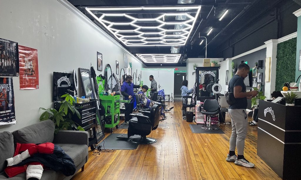 The inside of groom culture. Hardwood floors lead back to barber chairs on each side there are white neon honeycomb lights above and a black ceiling.