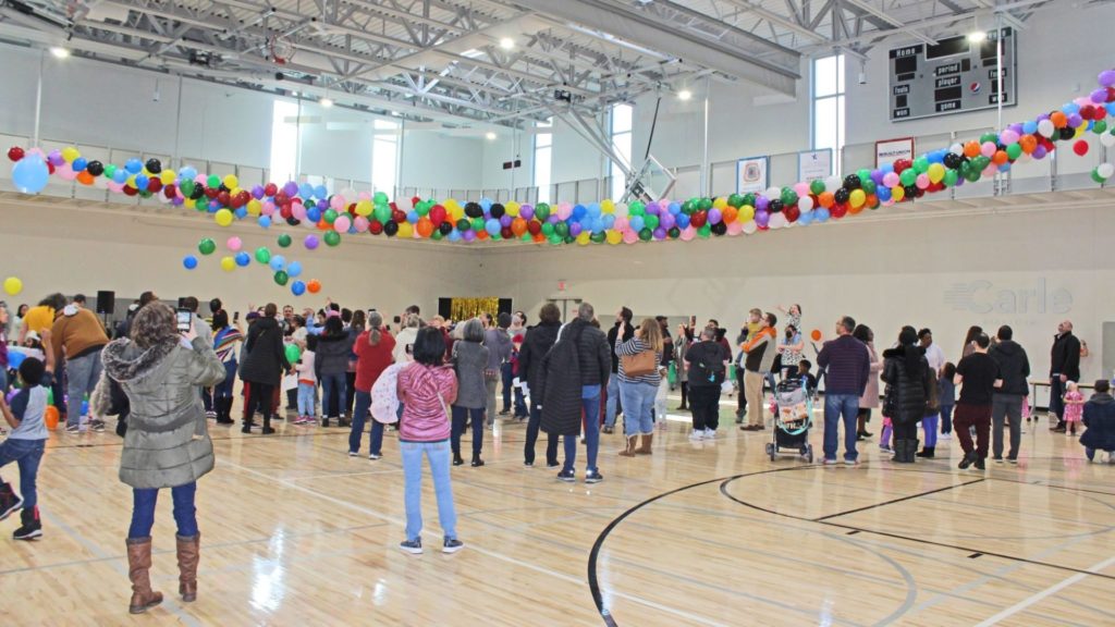 A group of adults and children gathered on a gym floor, watching colorful balloons falling from the ceiling.