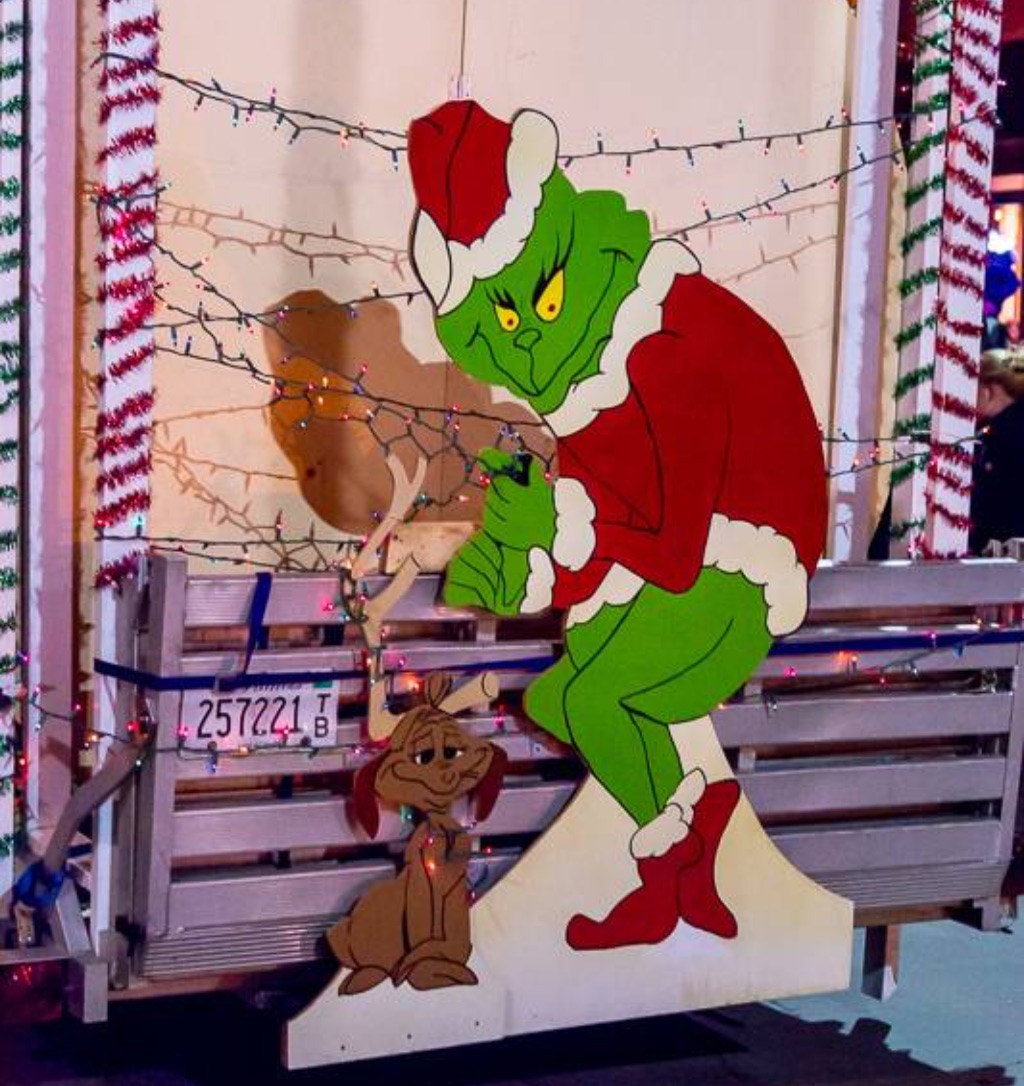 a cut out of the grinch standing in front of a white truck with Christmas lights on it.