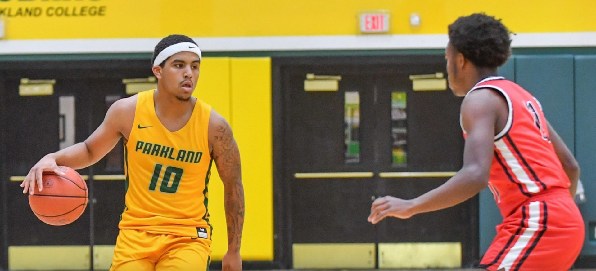 Parkland College men's basketball player Brylan Phillips, a Black man, wears a yellow Parkland uniform and a white nike sweatband. He is dribbling a basketball. A player from the opponent's team, a Black man wearing a red uniform, guards him. They are in a gym with yellow walls.