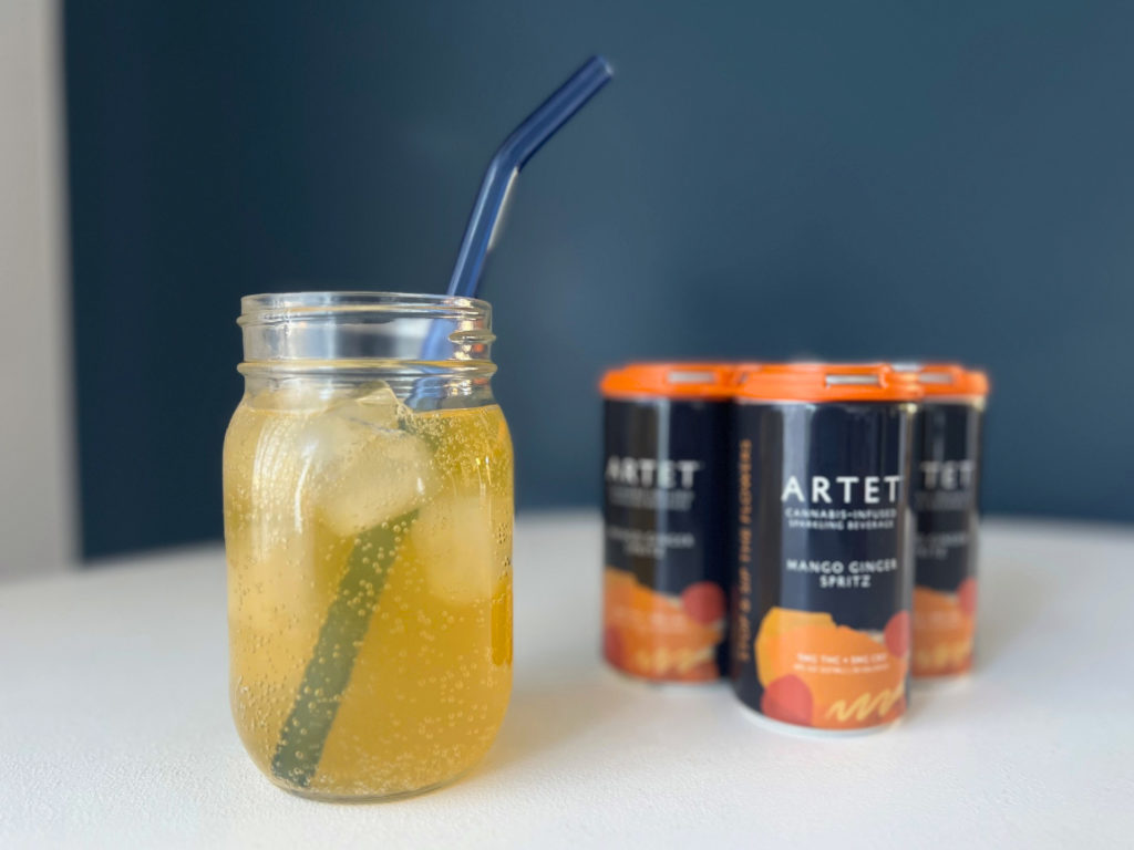 A mason jar of yellow-orange liquid on ice beside a four pack of Artet mango ginger spritz.