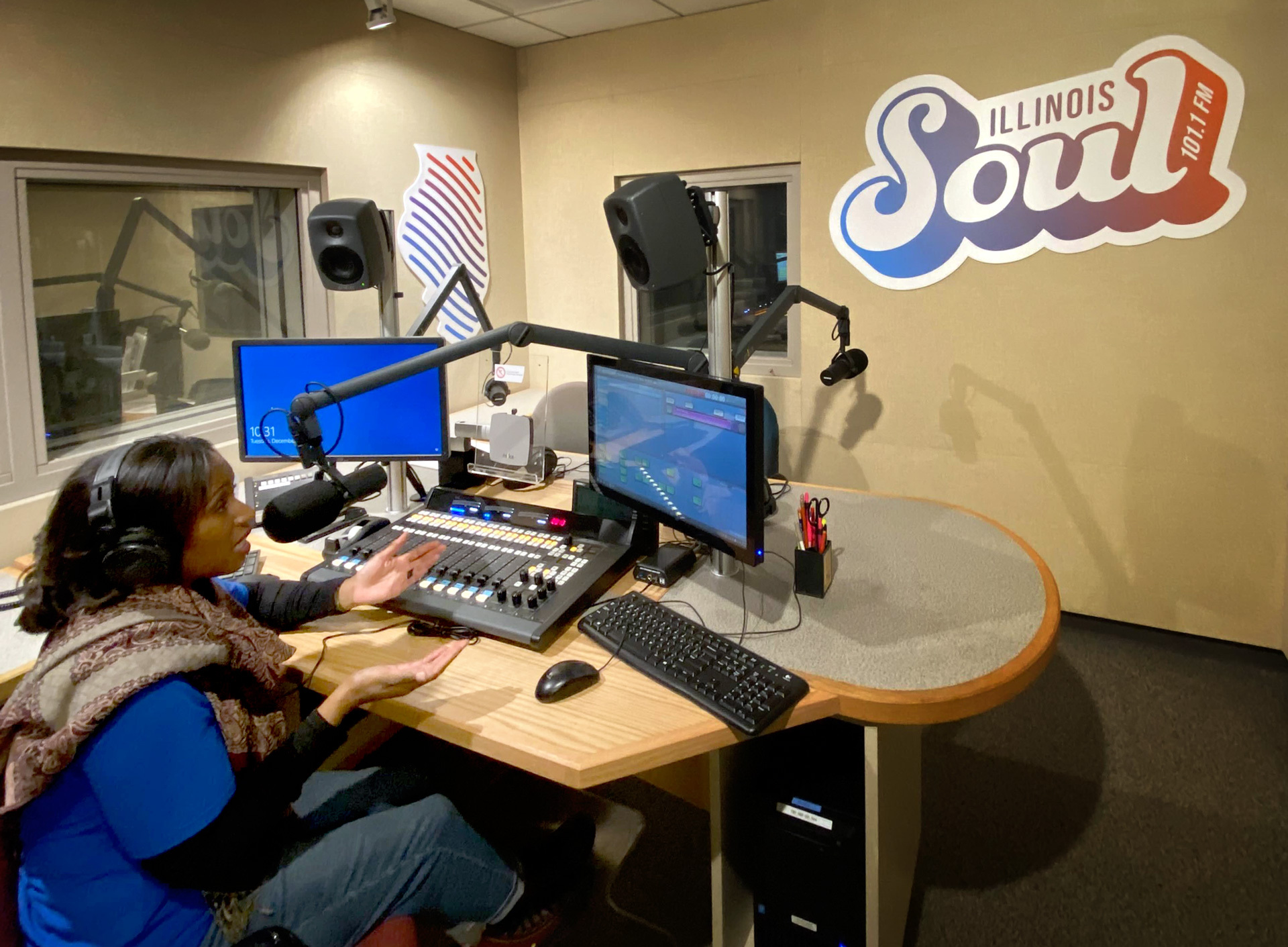 A Black woman sits behind a microphone in a radio studio, with two computer moniters is front of her. There is a sign that says Illinois Soul on the wall.
