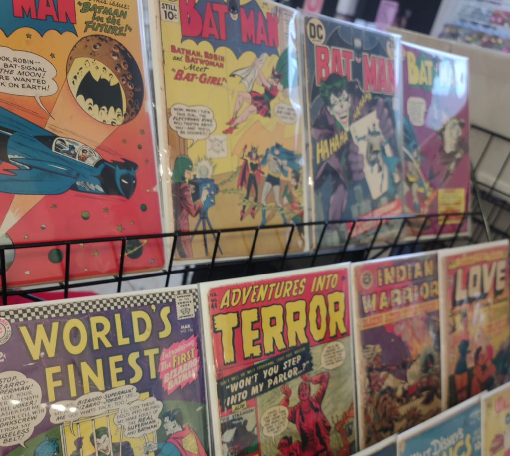 A black metal wire shelf holding three rows of vintage comic books in clear plastic sleeves. The first row is a collection of Batman books; the second row is varied, and the third row shows Disney comics.