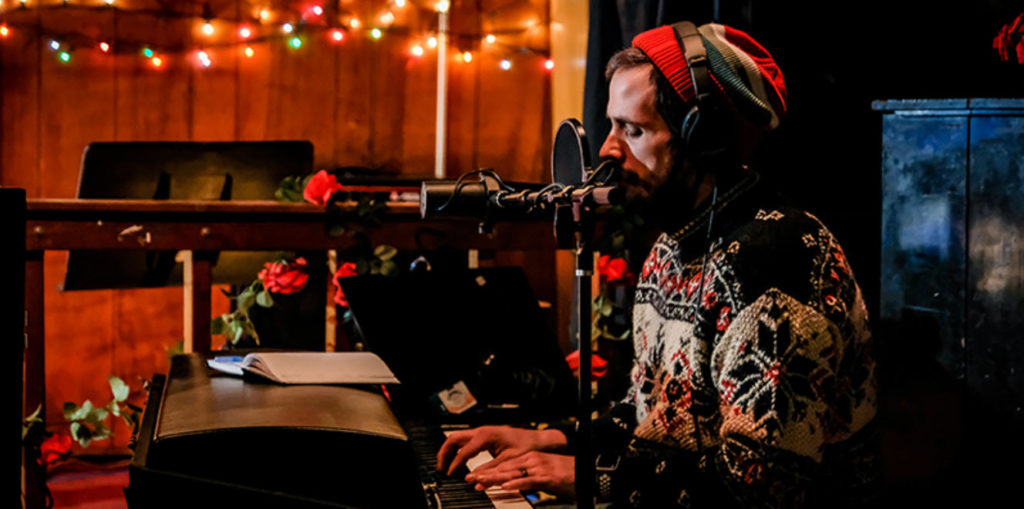 The setting is indoors with wooden walls adorned with string lights that cast a warm glow. A person is playing the piano; their attire consists of a patterned sweater and a beanie. The piano is black, and sheet music rests upon it. There are red roses intertwined with green leaves decorating the area around the piano. A microphone on a stand is positioned near the player. The overall ambiance is cozy and inviting, with a sense of warmth emanating from the scene. The person’s posture suggests they are deeply engaged in playing the piano. The patterned sweater and beanie give a casual and comfortable appearance. The roses add a touch of elegance and color to the scene. The microphone suggests a performance or recording might be taking place. The string lights and wooden walls contribute to a rustic and homely atmosphere. The image captures a moment of music and creativity in a charming and intimate setting.