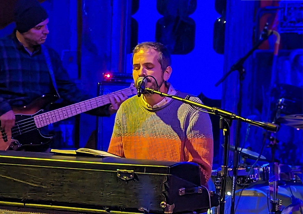 Two musicians are immersed in their performance on a stage bathed in blue light. The musician on the left is strumming a black electric bass guitar, dressed in a dark-colored long-sleeve shirt. The musician in the center is playing a keyboard, donned in a vibrant striped sweater with hues of orange, yellow, and white. A microphone stand extends across the front of the keyboard, and various musical equipment like speakers can be seen in the background. Their passion for music is palpable in this captivating scene.