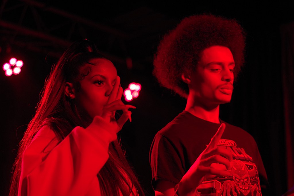 Two individuals are under the glow of intense red lighting, possibly in a stage or club setting. The individual on the left, with long hair, is wearing a white top and appears to be speaking into a microphone. The individual on the right, sporting an afro hairstyle, is dressed in a dark t-shirt with a visible design and is making a gesture with their hand. The overall atmosphere is vibrant, suggesting a lively performance or event.