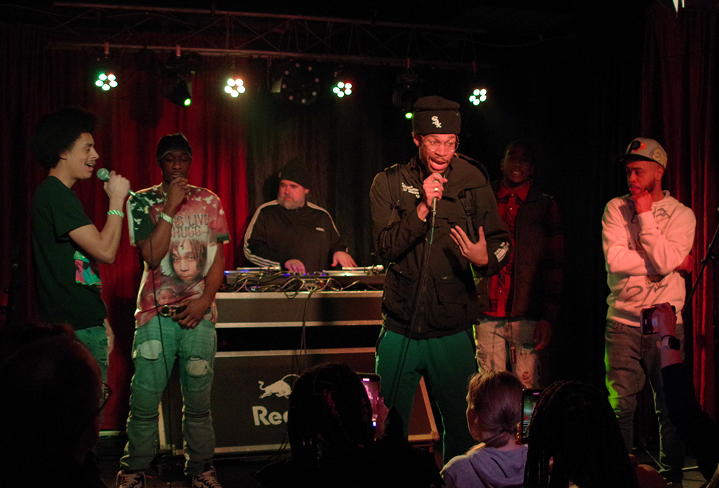 Six individuals are on a stage, bathed in overhead green lighting. The person on the far left is wearing a green t-shirt and jeans, holding a microphone close to their mouth. Next to them, another individual is in a graphic tee and ripped jeans. In the center, someone is behind a DJ booth, wearing dark clothing. The fourth person is dressed in a black jacket and green pants, also holding a microphone. The last individual on the right wears a white hoodie and light-colored jeans. The overall atmosphere suggests a lively performance or event. The text “Red Bull” is visible, indicating a possible sponsorship or event organizer. The image conveys a sense of energy and excitement.