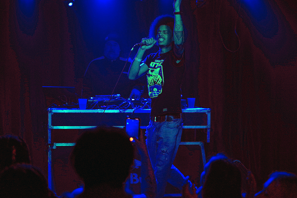 Under the glow of blue lighting, a person stands on stage, holding a microphone in one hand and raising the other. They are dressed in a graphic t-shirt and jeans, with tattoos visible on their raised arm. Behind them, another individual operates electronic equipment from a DJ booth. The audience’s silhouettes are visible in the foreground, and red curtains drape the background, suggesting a lively performance or event. The overall atmosphere is vibrant and dynamic.