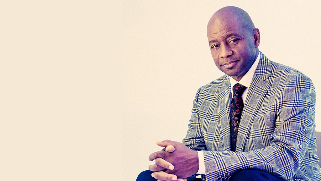 Branford Marsalis, a Black man is seated, wearing a well-tailored, grey plaid suit with a white shirt underneath. They are also wearing a dark tie that features an intricate red pattern.Their hands are clasped together in front of them. The background is plain and light-colored, putting focus on the individual. The individual’s arms are crossed or hands together, suggesting they might be in mid-conversation or contemplation.
